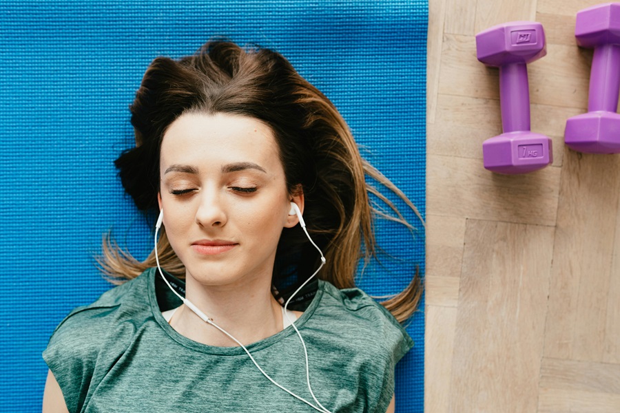 10 Songs to Listen to When you Want to Make a Deal a Woman Laying on a Yoga Mat with Earbuds in Her Ears and Two Dumbbells on the Ground Next to Her