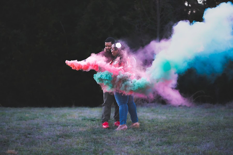 DIY Gender Reveal Ideas a Couple Standing Together Holding Smoke Bombs with Pink and Blue Smoke Flowing Around Them