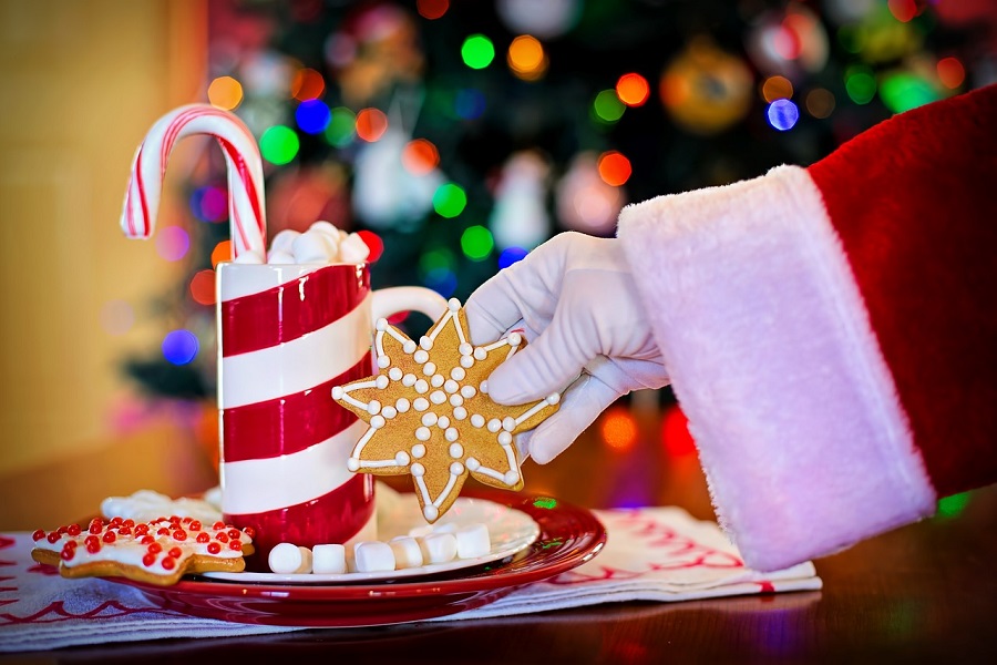 Things to Do on Christmas Morning Close Up of Santa's Hand Reaching for a Cookie on a Plate Next to a Red and White Striped Cup of Hot Chocolate Garnished with a Candy Cane