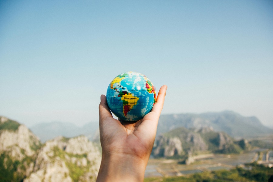10 Travel Quotes for the Wanderlust a Person's Hand Held Outward Holding a Small Toy Earth Globe with Mountains in the Background
