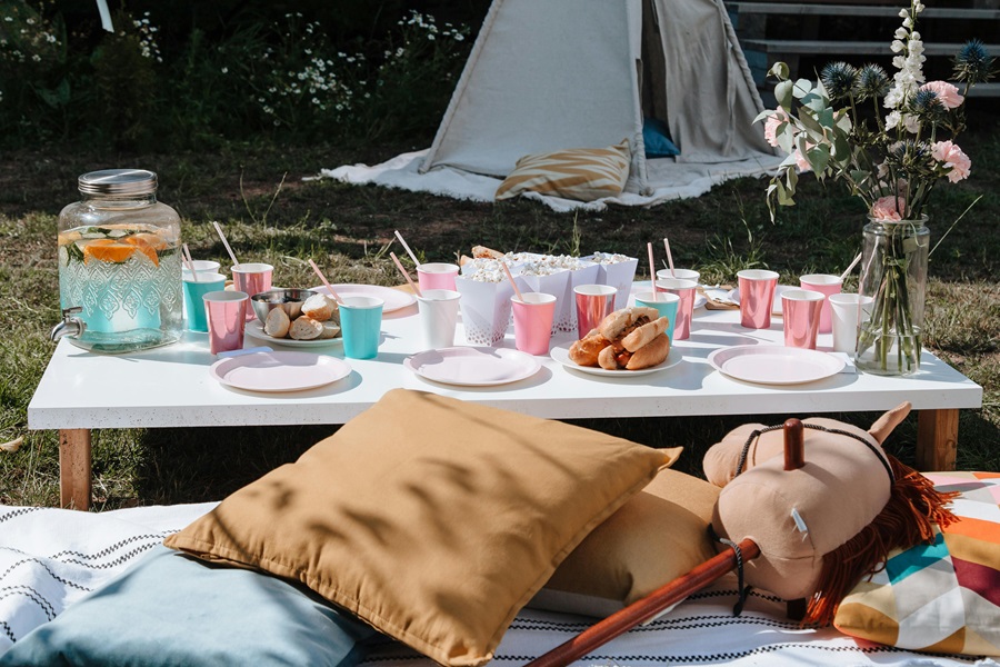 Summer Party Essentials Shopping List View of a Table with Plates, Cups, and Silverware Outside with a Tent in the Background and Pillows in the Foreground