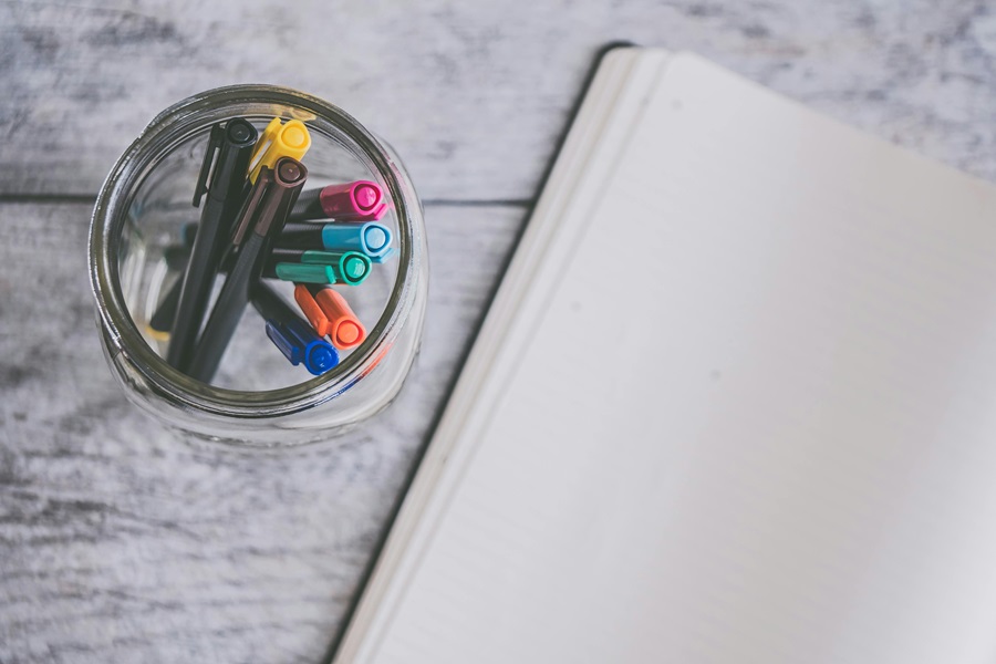 Bullet Journal Work Ideas to Get Your Hustle On an Open Bullet Journal with a Glass Cup of Colored Pens Next to it on a Table