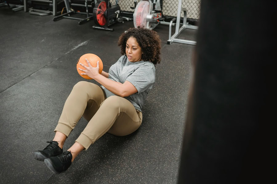 Muffin Top Workouts a Woman Sitting on the Ground Holding a Medicine ball in Her Hands to One Side