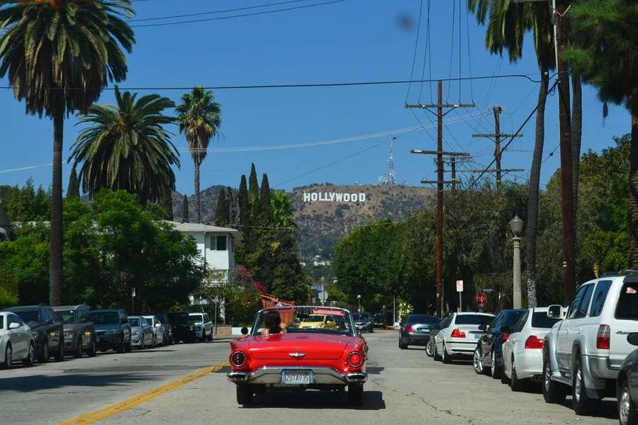 Best Tourist Things to Do in LA a Classic Red Car Driving Down a Road Toward the Hill with the Hollywood Sign on Top