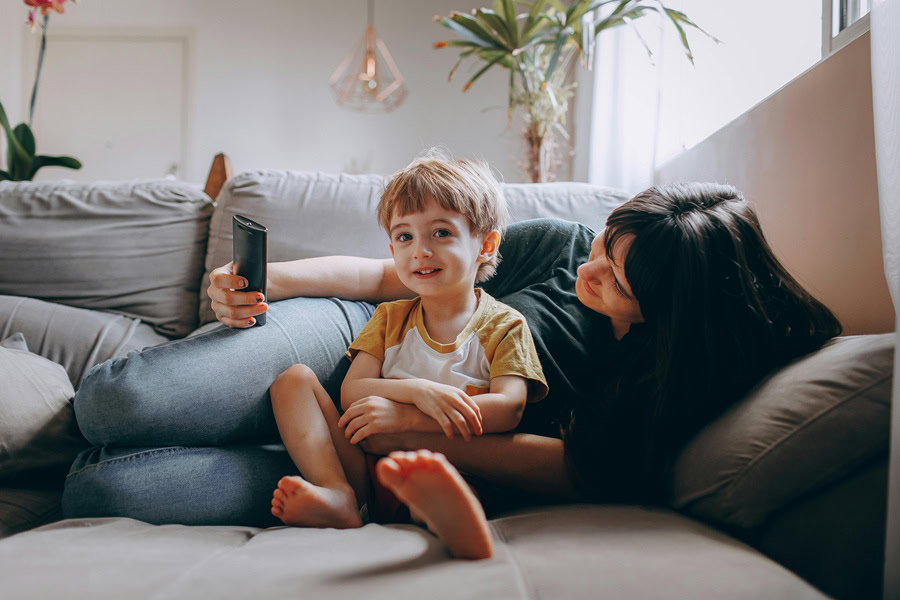 Family Friendly Movies to Watch at Home a Mom and Her Son Laying on a Couch Holding a TV Remote