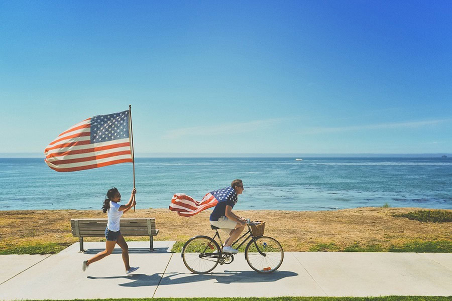 10 Fun 4th of July Instagram Captions Two People Outside with American Flags, One Riding a Bike and the Other Running Behind Them