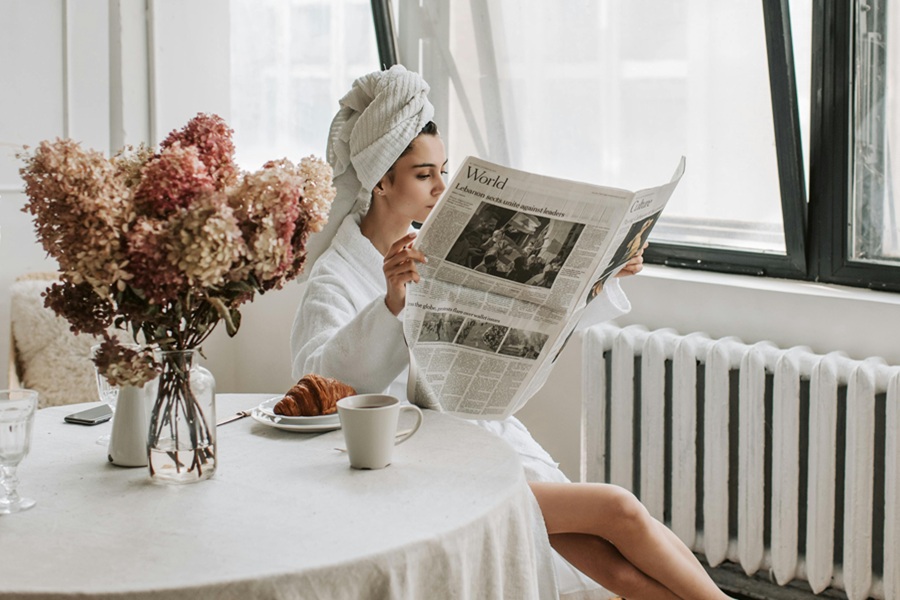 How to Start Your Day a Woman Sitting at a Kitchen Table Wearing a Towel Wrapped Around Her Hair Reading a Newspaper with an Open Window in the Background