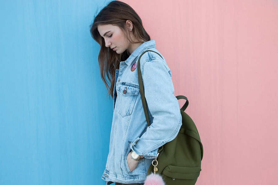 Ways to Wear a Jean Jacket a Woman Wearing a Jean Jacket and a Backpack Standing Next to a Wall That is Painted Half Blue and Half Pink