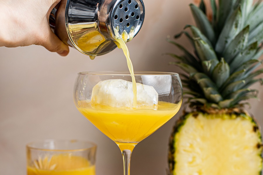 Banana Creme Rum and Orange Juice Cocktail Someone Pouring the Cocktail from the Shaker into a Glass with a Pineapple Cut in Half in the Background