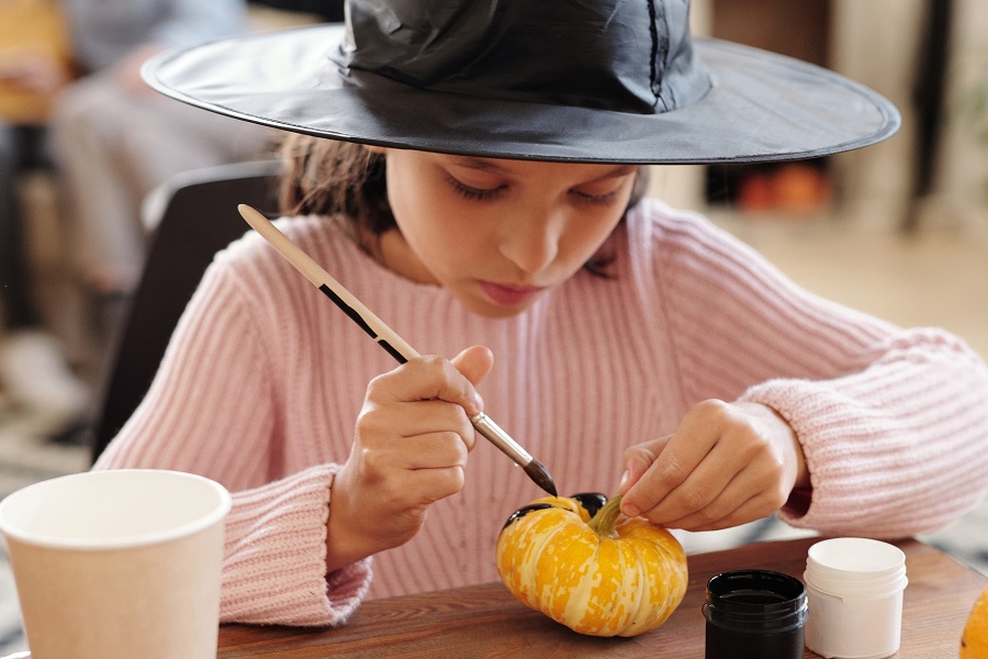 DIY No Carve Pumpkin Decorating Ideas Close Up of a Young Girl Wearing a Witch Hat Painting a Small Pumpkin