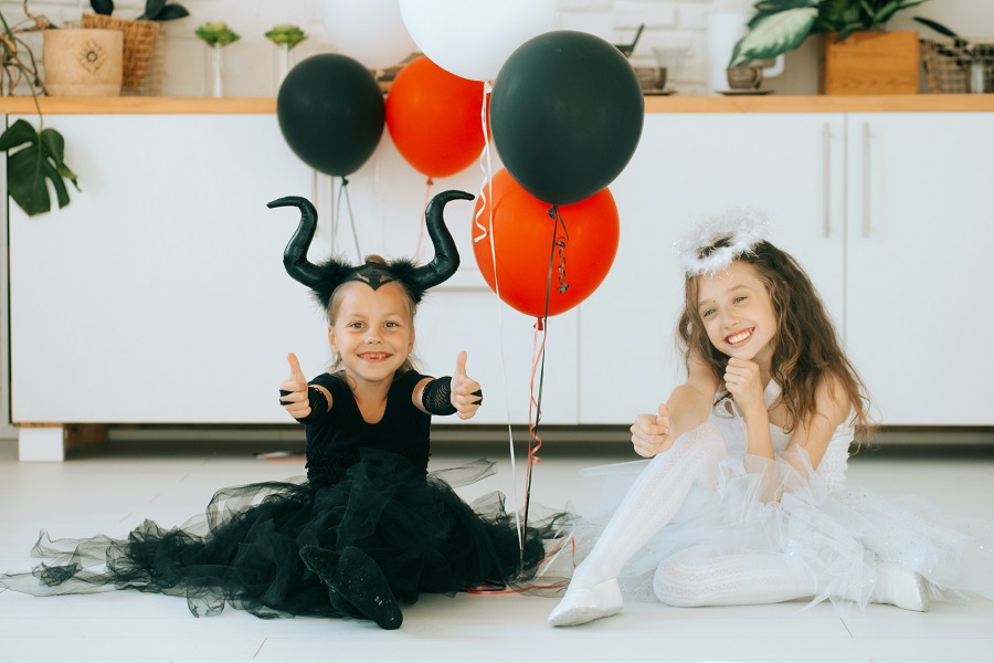 Halloween Games for Kids Two Little Girls Dressed up in Halloween Costumes Sitting on a Floor Smiling with Halloween Balloons Behind Them