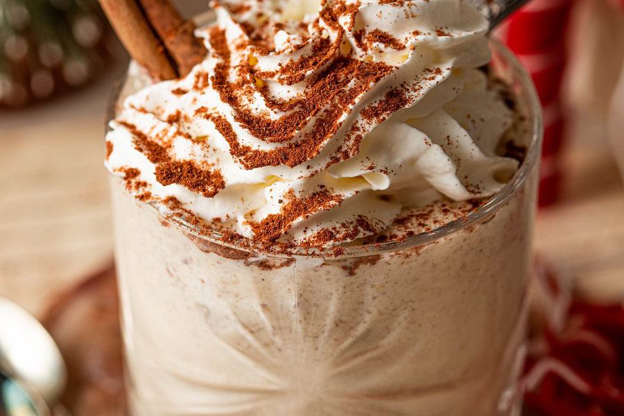 Bourbon Eggnog Recipe Close Up of a Glass of Eggnog Topped with Whipped Cream and Cinnamon with a Cinnamon Stick Sticking Out of the Cream