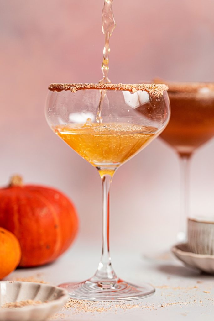 Pumpkin Spice Mocktail Mixture Being Poured into a Glass with Graham Cracker on the Rim and Pumpkins in the Background