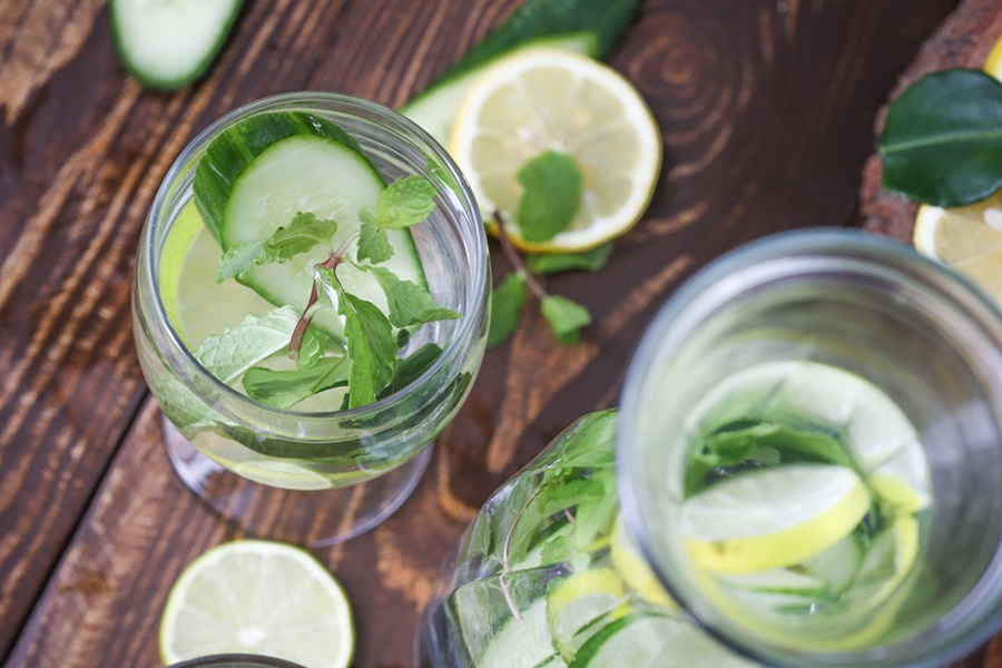 Detox Water Recipes for Weight Loss Overhead View of a Pitcher of Water Next to a Glass of Water Both with Herbs and Slices of Lemon and Cucumber in Them