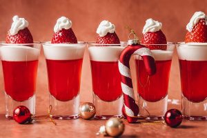 Easy Santa Hat Jello Shot Recipe Close Up of a Row of Jello Shots with a Candy Cane Tree Ornament Leaning Against Two of the Shot Glasses