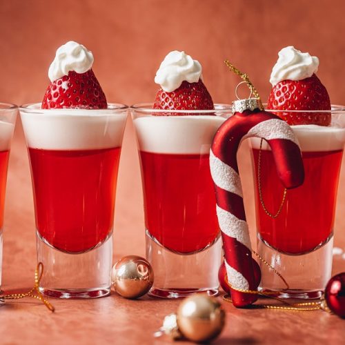 Easy Santa Hat Jello Shot Recipe Close Up of a Row of Jello Shots with a Candy Cane Tree Ornament Leaning Against Two of the Shot Glasses