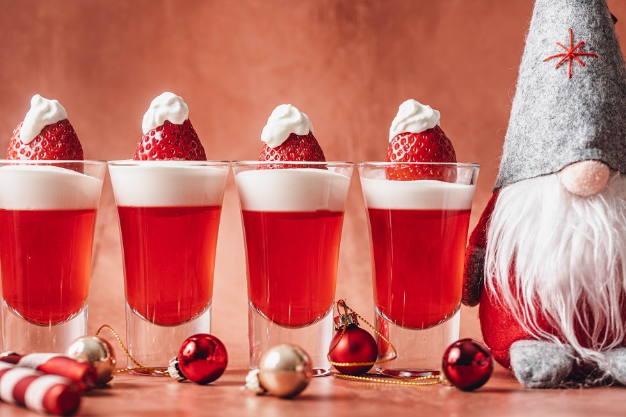 Easy Santa Hat Jello Shot Recipe a Row of Jello Shots with a Small Gnome Dressed Like an Elf Leaning Next to Them