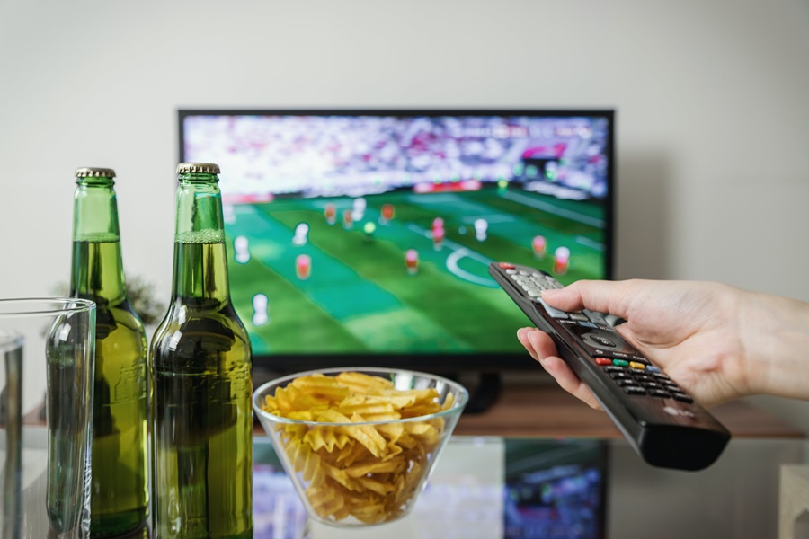 Super Bowl Party Food a Person's Hand Pointing a Remote at a TV with Football On and Chips and Beer on a Coffee Table Between the Person and the TV