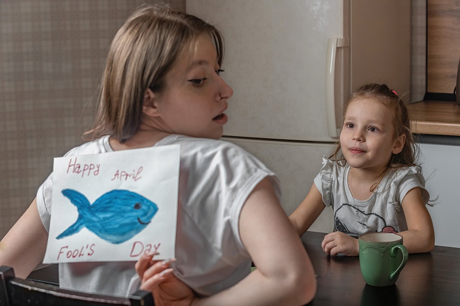 April Fools Day Quotes Mom Reaching Behind Her Back To Take Off a Piece of Paper with a Fish Drawn on It and a Little Girl Sitting in Front of Her Smiling