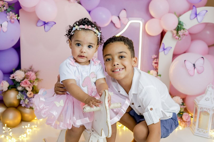 1st Birthday Party Ideas for Girls a Brother Posing with His Baby Sister in Front of a Pink and Purple Backdrop with a White "1" on the Wall