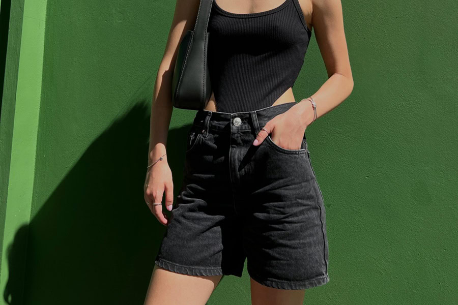 Women's Jort Outfit Ideas a Woman Wearing a Black Bathing Suit with Black Jorts and a Black Purse Leaning Against a Green Wall