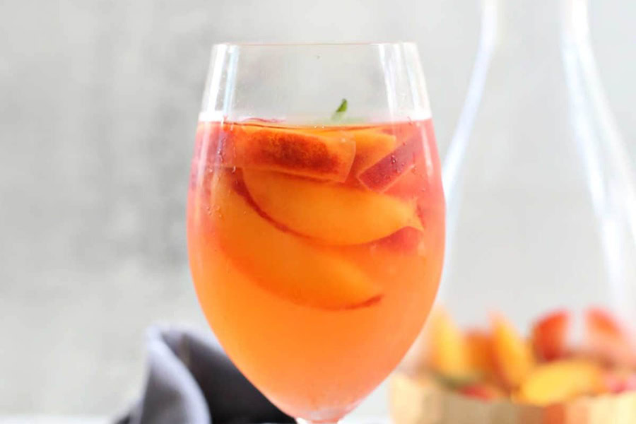 Sangria Recipes White Peach Sangria in a Wine Glass with a Pitcher Filled with Peach Slices in the Background