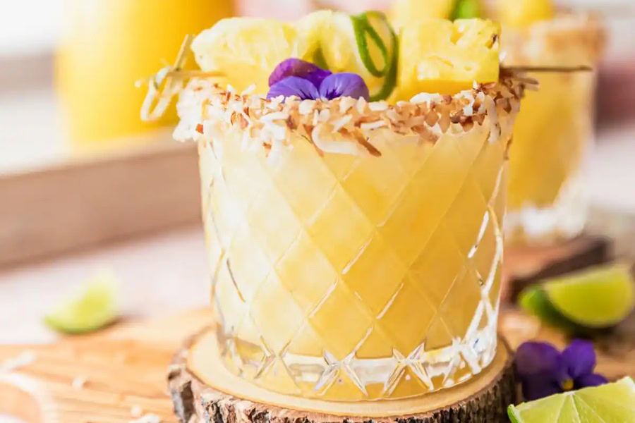 Backyard BBQ Drinks for an Outdoor Party Creamy Pineapple Coconut Margaritas in a Glass Garnished with Pineapple Wedges and Edible Flowers