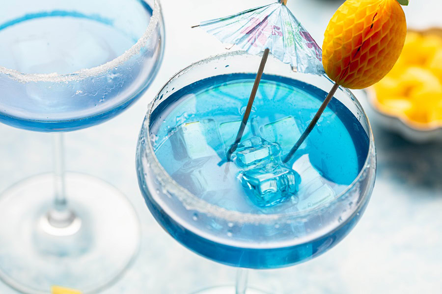 Backyard BBQ Drinks for an Outdoor Party Blue Margaritas Pitcher Recipe in a Glass with a Small Umbrella Toothpick and a Small Paper Pineapple on a Stick